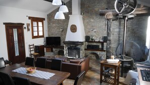 Un salon chaleureux avec une cheminée en pierre, un coin repas avec une table et des chaises, et une cuisine équipée d’une cuisinière.