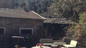 Une terrasse extérieure avec des tables en pierre et des chaises en osier, entourée d’un bâtiment en pierre et offrant une vue sur une colline, avec, au loin, une structure ressemblant à un château.
