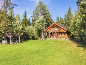 A wooden cabin with a porch, surrounded by a grassy yard and tall trees.