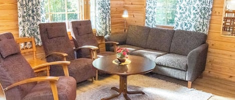 A cozy log cabin interior with a sofa, two armchairs, a small round table with a vase, and a lamp.
