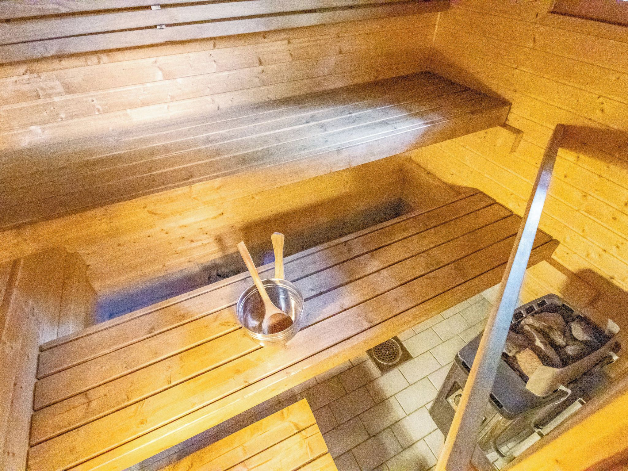Ein Saunaraum mit Holzbänken, einer Sitzbank mit Metallbecken und Holzlöffel sowie einem gefliesten Boden.