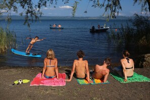A group of people sitting on beach towels by a lake, with a boat and another person on a surfboard in the background.