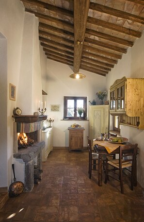 A rustic kitchen with a stone fireplace, wooden beams, and a dining area with wooden chairs and a table.