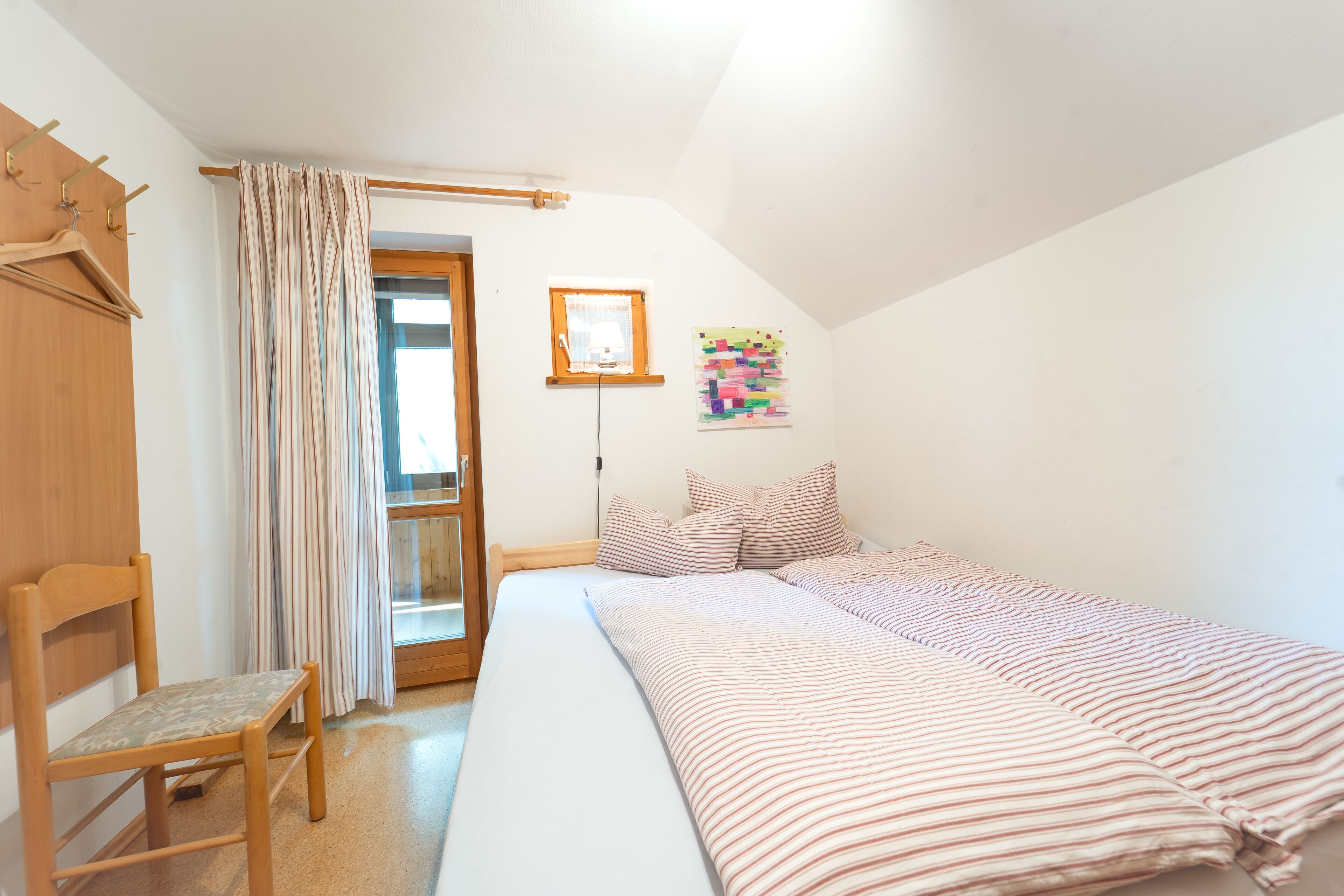 Un dormitorio con una cama, una silla de madera, una ventana con cortinas, un cuadro en la pared y una puerta.