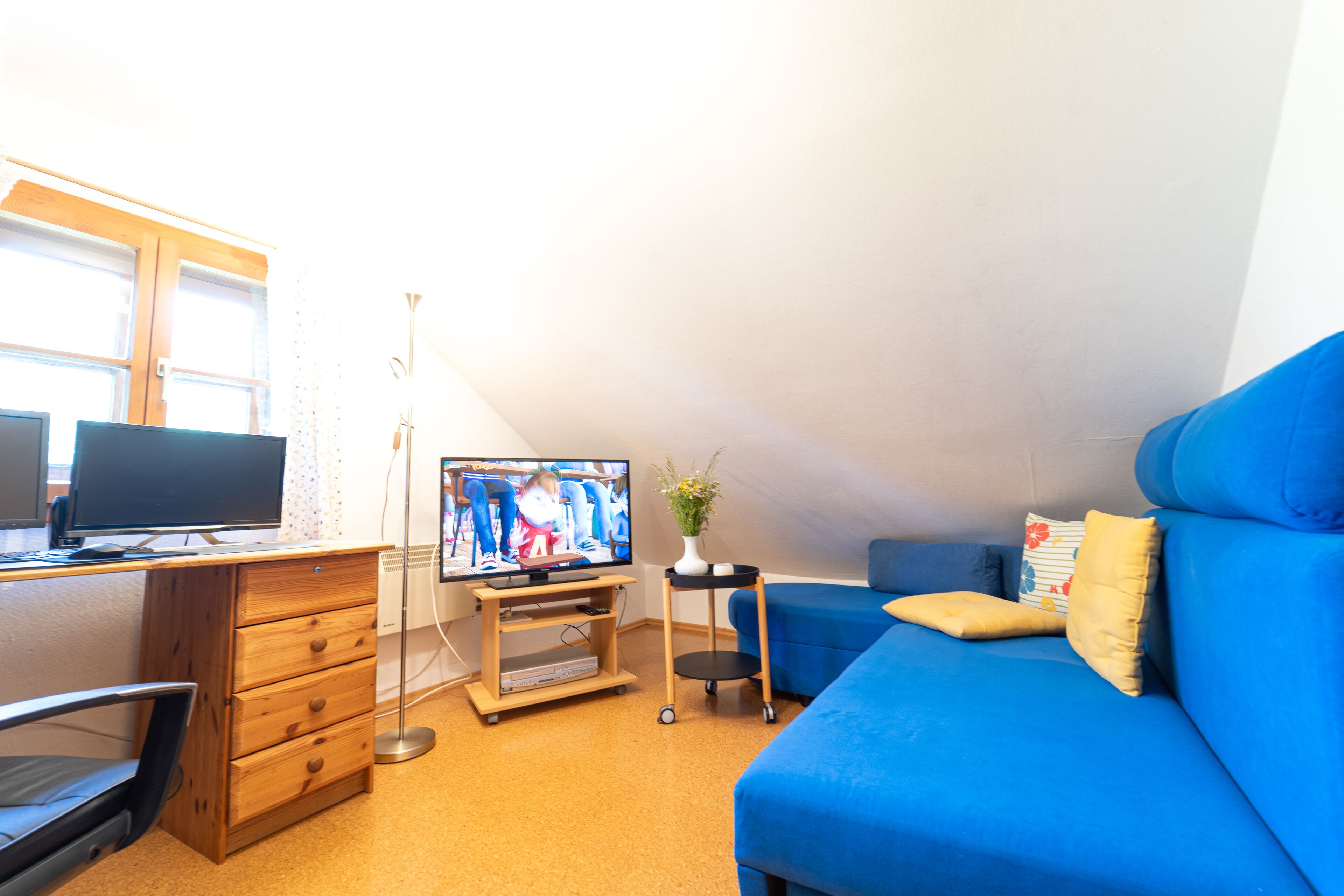 Una habitación pequeña y luminosa con un escritorio de madera, un televisor, un sofá azul y una mesita con un florero.