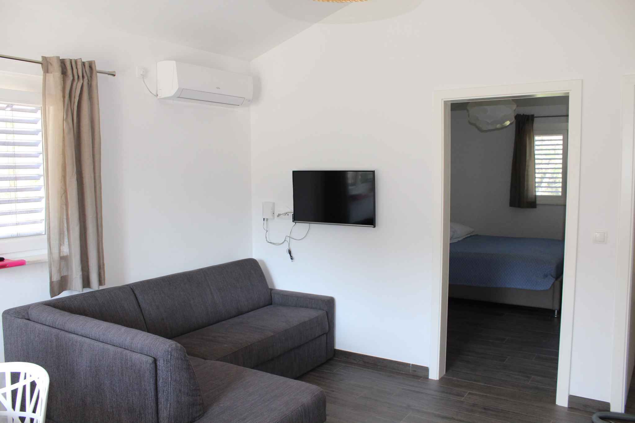 A room with a grey sofa, a flat-screen TV mounted on the wall, a bed with a blue bedspread, and an air conditioning unit.