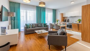 Ein modernes Wohnzimmer mit Sofa, Sessel, Couchtisch und einem Essbereich mit Stühlen und Tisch. Im Hintergrund ist eine Küche mit weißen Schränken und einem Herd zu sehen.