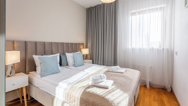 Ein modernes Hotelzimmer mit einem großen Bett, zwei Nachttischen mit Lampen, einem grauen Kopfteil und einem großen Fenster mit durchsichtigen Vorhängen.