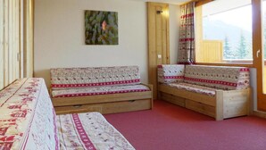 A room with a wooden sofa set, a bed with patterned bedding, a painting on the wall, and a window with a view of trees and mountains. - 10m from ski slopes, 1st floor, balcony, 27m², Méribel-Mottaret (Méribel - Les Allues)
