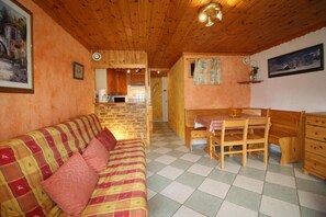 A cozy room with a wooden ceiling, a checkered floor, a sofa with a plaid blanket, a wooden dining table with chairs, and a kitchen area visible through an open door.
