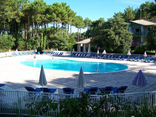 A pool area with blue lounge chairs and umbrellas, surrounded by trees and a building with a balcony.