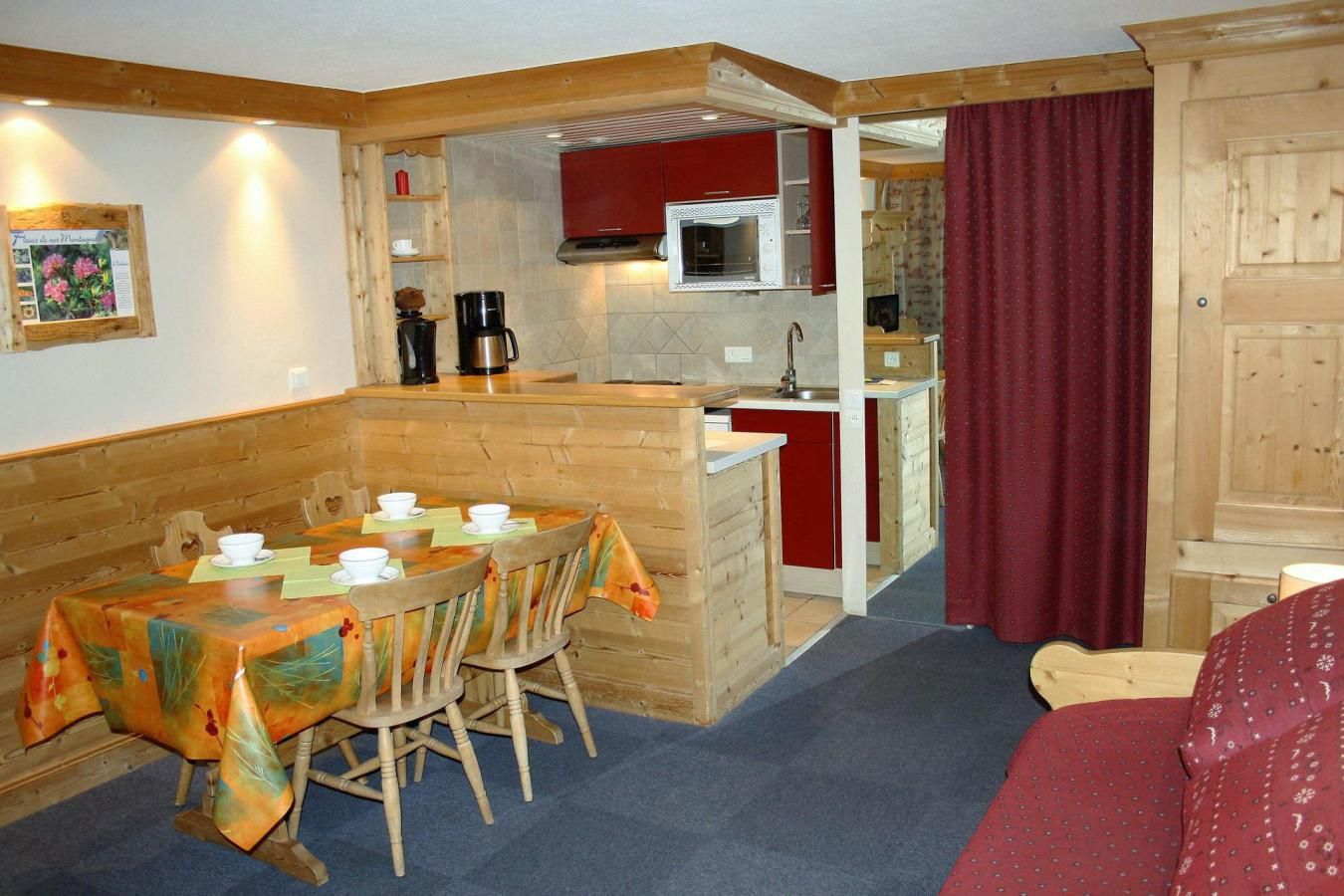 A cozy room with a wooden dining table set for four, a kitchen area with red cabinets, and a large wooden wardrobe.