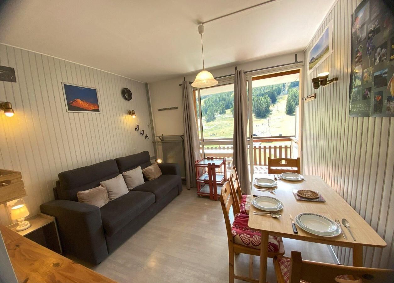 A cozy room with a sofa, dining table set for four, and a view of a forest through a window.