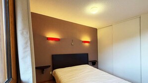 Une chambre à coucher avec un lit, une table de chevet, une lampe fixée au mur et une fenêtre avec des rideaux.
