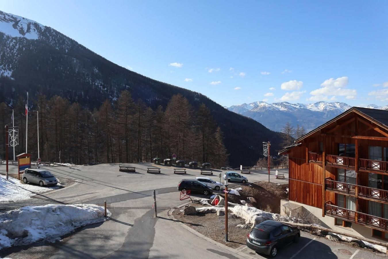 Un albergue de montaña con zona de estacionamiento, un edificio de madera con balcones y vistas a montañas nevadas.