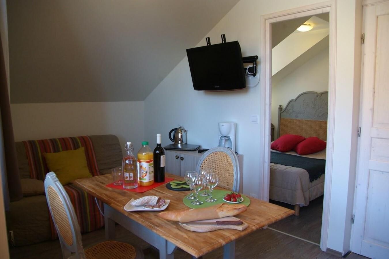 Ein Essbereich mit Holztisch, Stühlen, einem an der Wand befestigten Fernseher und einem Bett mit roten Kissen im Hintergrund.