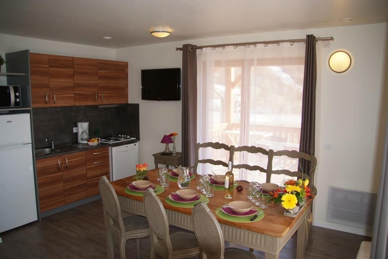 On aperçoit un coin repas avec une table en bois dressée pour quatre personnes, entourée de chaises. Une cuisine avec des meubles en bois et un four se trouve sur la gauche.