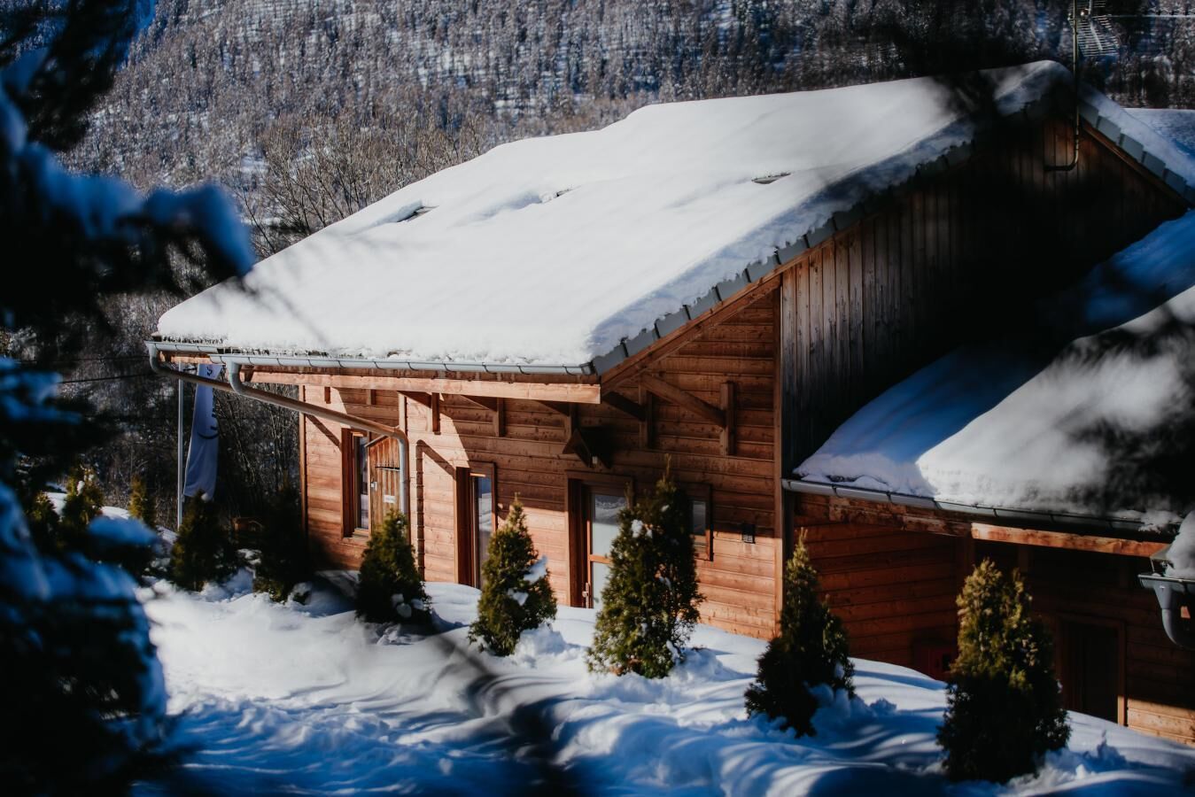 Uma cabana de madeira com telhado coberto de neve, cercada por árvores e um cenário montanhoso.