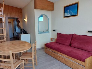 A compact living space with a wooden dining table and chairs, a red sofa, a small kitchenette, and a framed picture on the wall.