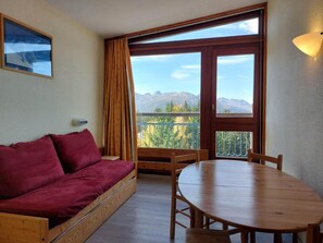 A room with a wooden sofa, a wooden table, and a balcony with a view of mountains.