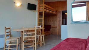 Een kamer met een stapelbed, een houten tafel met stoelen, een rode bank, een kleine televisie aan de muur en een raam met gordijnen.