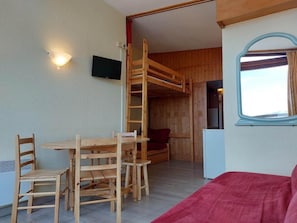 A room with a bunk bed, a wooden table with chairs, a red sofa, a small TV on the wall, and a window with curtains.
