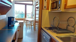 Een kleine keuken met een magnetron, koffiezetapparaat en een spoelbak. Achtergrond toont een eethoek met een tafel en stoelen.