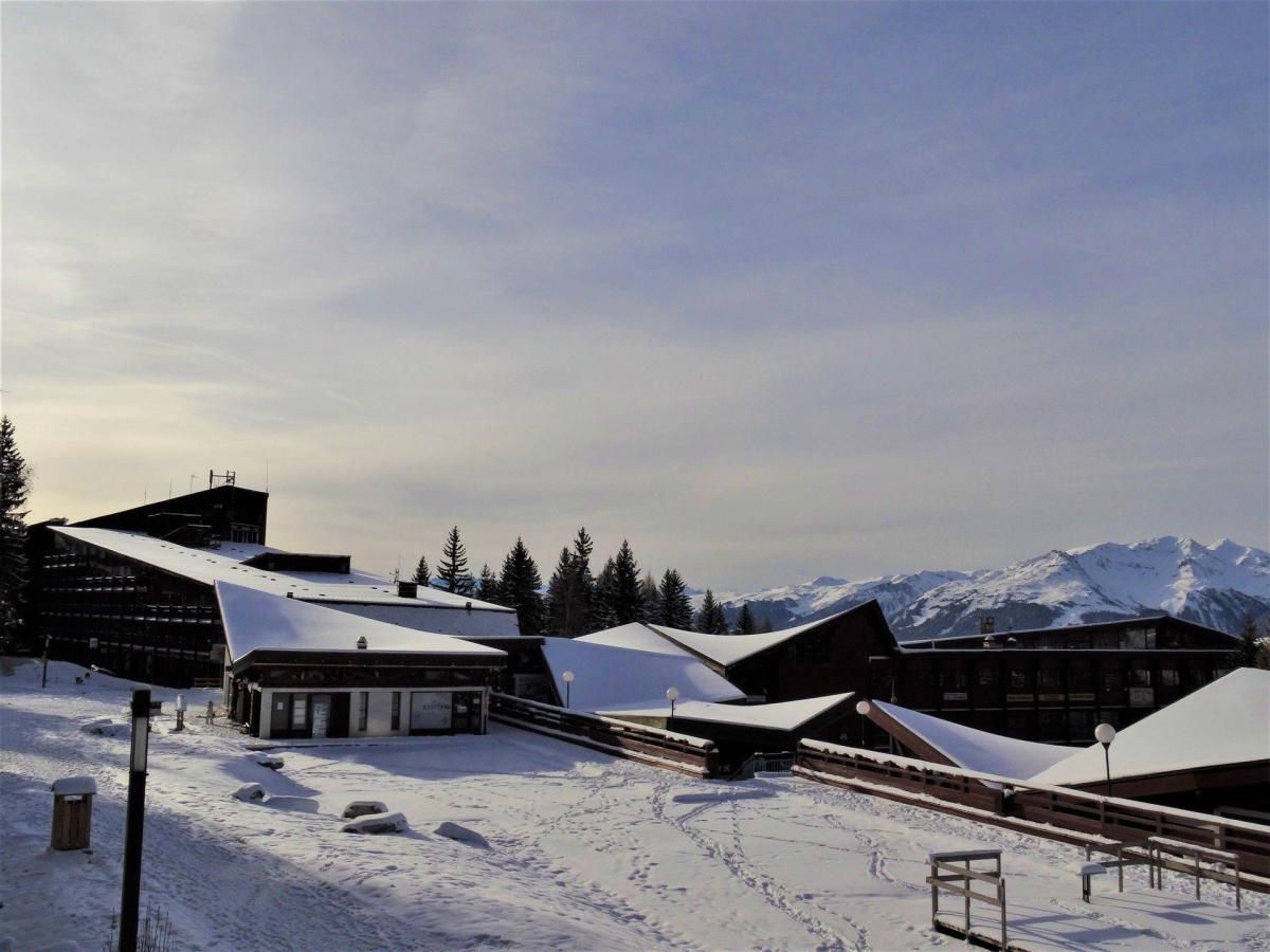 A snow-covered ski resort with modern buildings, a ski lift, and a mountain backdrop.