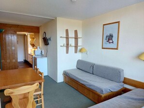 A compact room with a wooden dining table, a grey sofa, a small kitchenette, and a wooden cabinet.