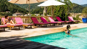A poolside area with lounge chairs and umbrellas, a pool, and a mountainous backdrop.