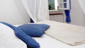 A bedroom with a bed, white curtains, a window with a view of greenery, and a white radiator.