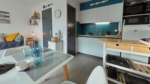 Una cocina compacta con electrodomésticos modernos, un comedor con mesa de vidrio y una zona de estar acogedora con sofá y cojines.