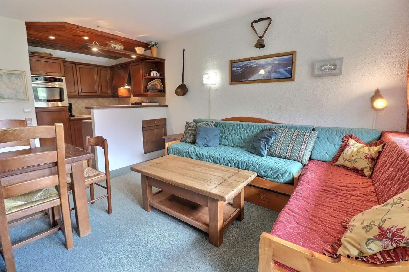 A cozy living area with a wooden dining table, a sofa with patterned cushions, and a kitchen in the background.