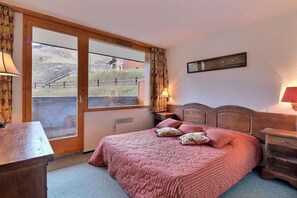A bedroom with a bed, a wooden headboard, a nightstand, a lamp, and a window with curtains. - 10m from ski slopes, Ground floor, balcony, 40m², Méribel-Mottaret (Méribel - Les Allues)