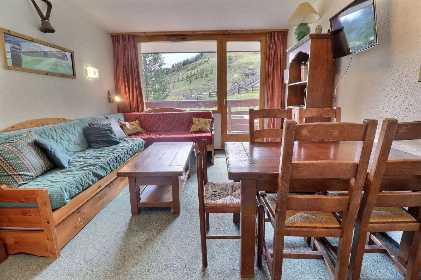 A cozy room with a wooden sofa, a dining table with chairs, a television, and a view of a hillside.