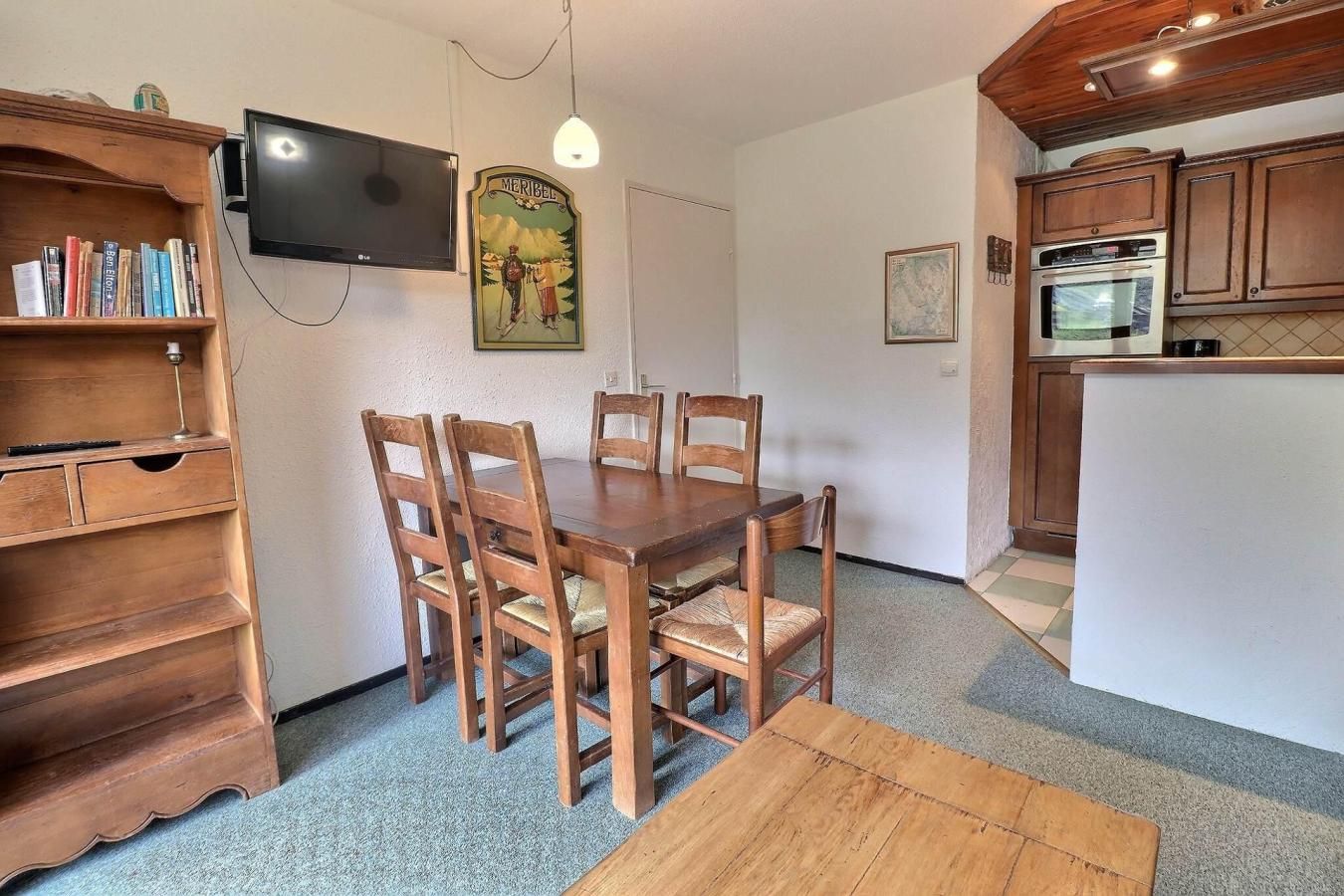 Uma sala de jantar com mesa e cadeiras de madeira, uma estante, uma televisão e uma cozinha com armários e eletrodomésticos de madeira.