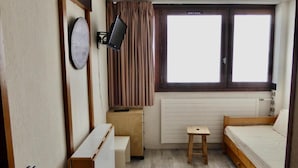 Une petite pièce, de taille modeste, comprenant un lit simple, un bureau, une chaise, un téléviseur fixé au mur et une fenêtre avec des rideaux.
