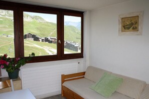 A room with a wooden bench, a radiator, a window with a view of a green landscape and buildings, and a framed picture on the wall.