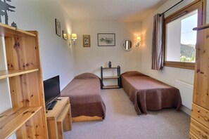 A small, cozy room with two single beds, a desk with a computer, and a window with curtains.