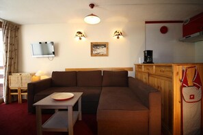 Uma sala de estar aconchegante com sofá, uma mesinha, televisão e um espaço de cozinha com cafeteira.