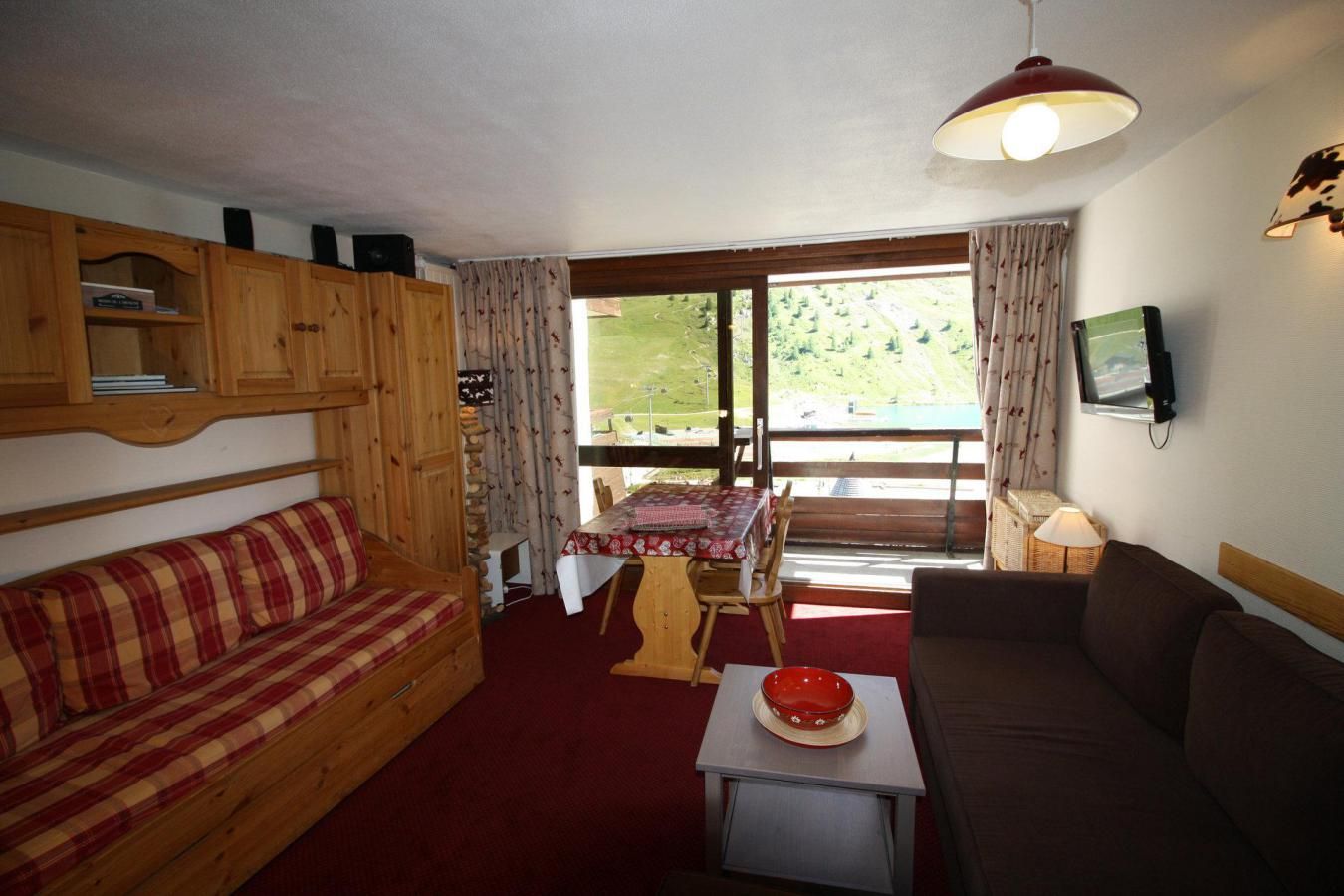 A cozy room with a wooden sofa, a red plaid sofa, a dining table with a red and white checkered tablecloth, and a television mounted on the wall.