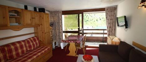 A cozy room with a wooden sofa, a red plaid sofa, a dining table with a red and white checkered tablecloth, and a television mounted on the wall.