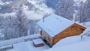 Uma cabana de madeira com telhado coberto de neve, rodeada por árvores e montanhas nevadas ao fundo.