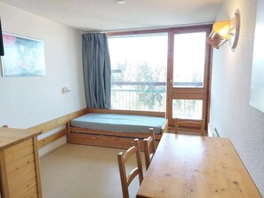 A small room with a wooden bed, a table, and a chair. There is a window with a curtain, a wall-mounted light, and a picture on the wall.