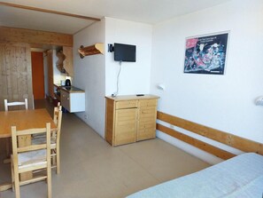 A room with a wooden dining table and chairs, a wooden wardrobe, a bed with a patterned bedspread, a mounted television, and a poster on the wall.