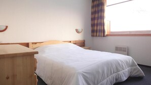 Un llit individual amb capçalera de fusta a una habitació amb finestra, una taula de nit i un mirall fixat a la paret.