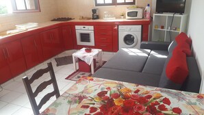Une cuisine compacte avec des meubles rouges, un four à micro-ondes, un lave-linge et un coin repas agrémenté d’une nappe à motifs floraux.