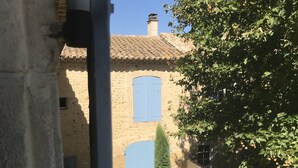 A rustic building with a tiled roof, two blue shutters, and a chimney. There are potted plants and a bench in the courtyard.