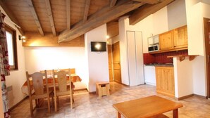 A cozy room with a wooden dining table, chairs, a small stool, a television mounted on the wall, and a kitchen area with wooden cabinets and a microwave. - 10m from ski slopes, 4th floor, balcony, tv, ski locker, 33m², Val Cenis (Lanslevillard)