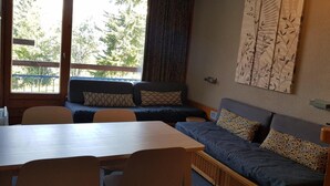 Una habitación acogedora con una mesa de madera, dos sofás con cojines estampados y una ventana con vistas a árboles.